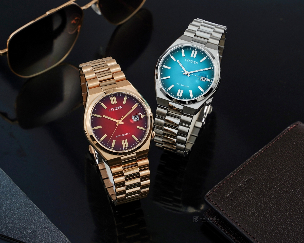Hai chiếc đồng hồ Citizen dạ quang dòng Tsuyosa Automatic đặt cạnh kính mát và ví da trên nền đen.