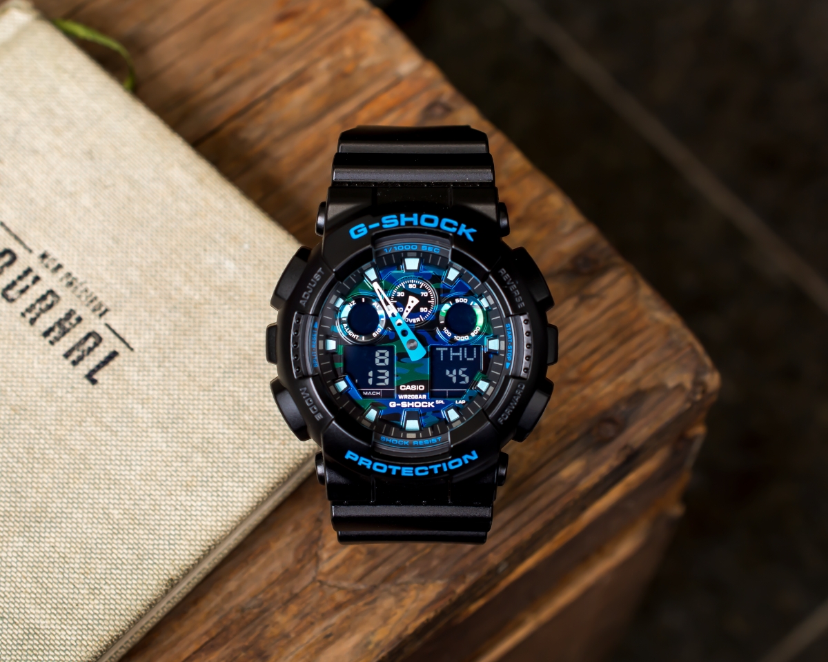 Đồng hồ G-Shock GA-100CB-1ADR phối xanh đặt trên bàn gỗ cạnh túi vải.