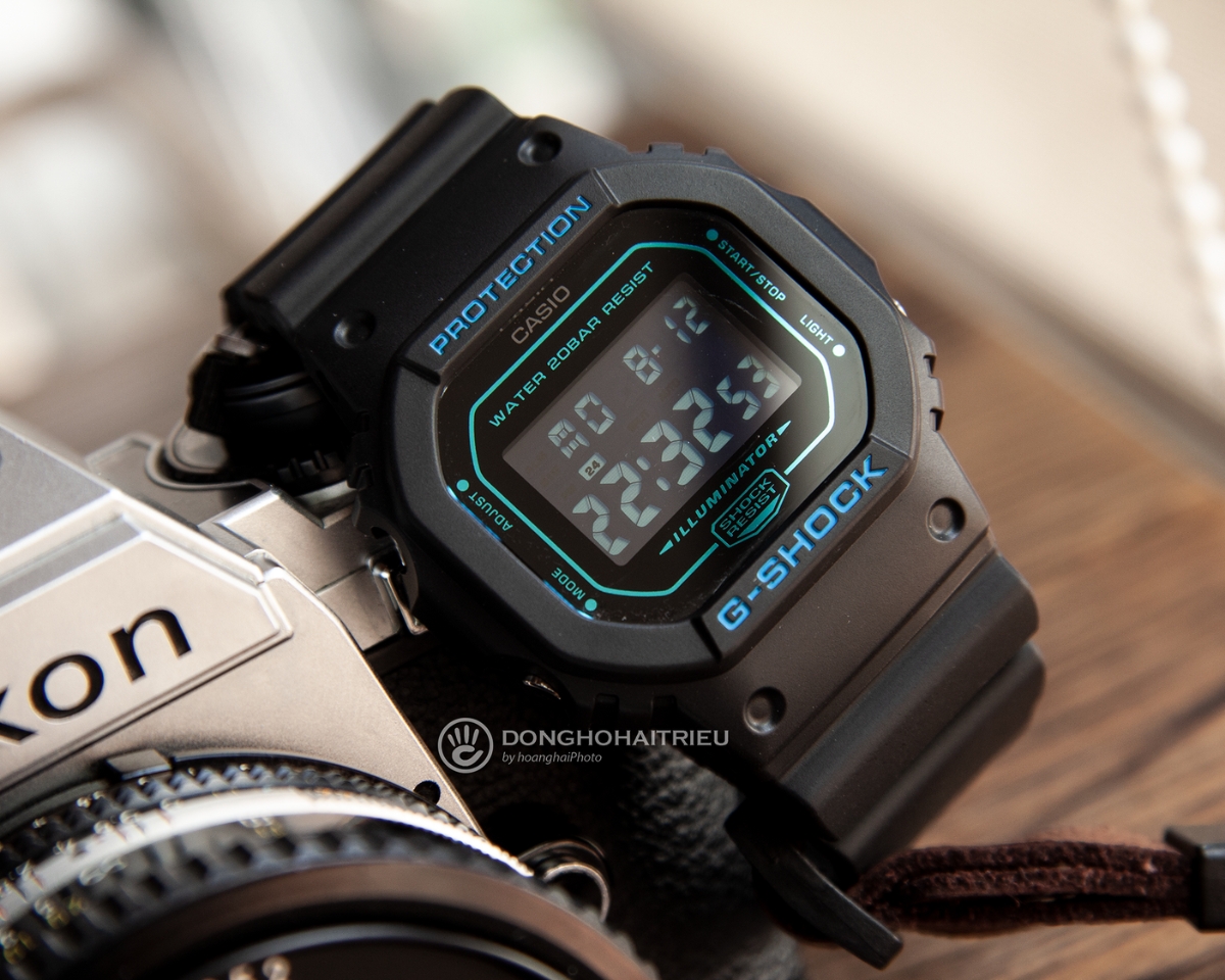 Đồng hồ G-Shock DW-5600BBM-1DR điện tử mặt vuông màu đen xanh đặt cạnh máy ảnh.