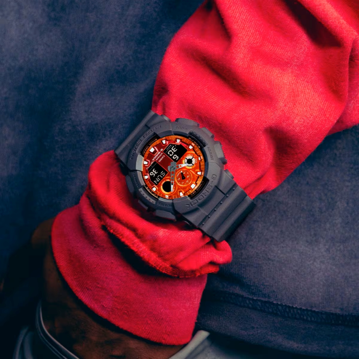 Đồng hồ G-Shock mặt cam đeo trên cổ tay với trang phục xanh đỏ.