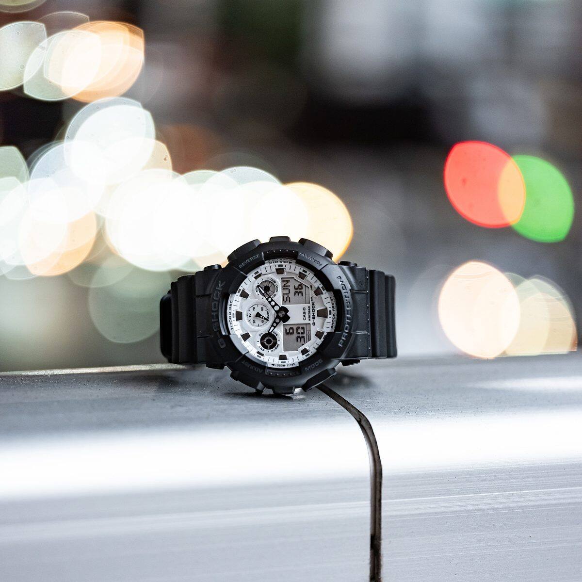 Đồng hồ G-Shock GA-100 mặt trắng đặt trên lan can với nền đèn thành phố.