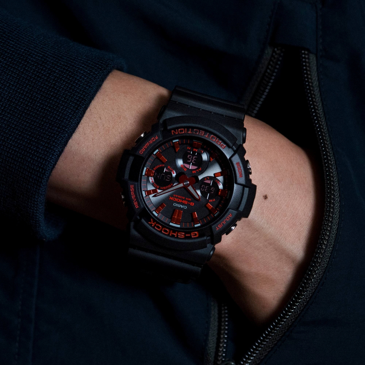 Đồng hồ G-Shock đen đỏ đeo trên cổ tay trong trang phục tối màu.