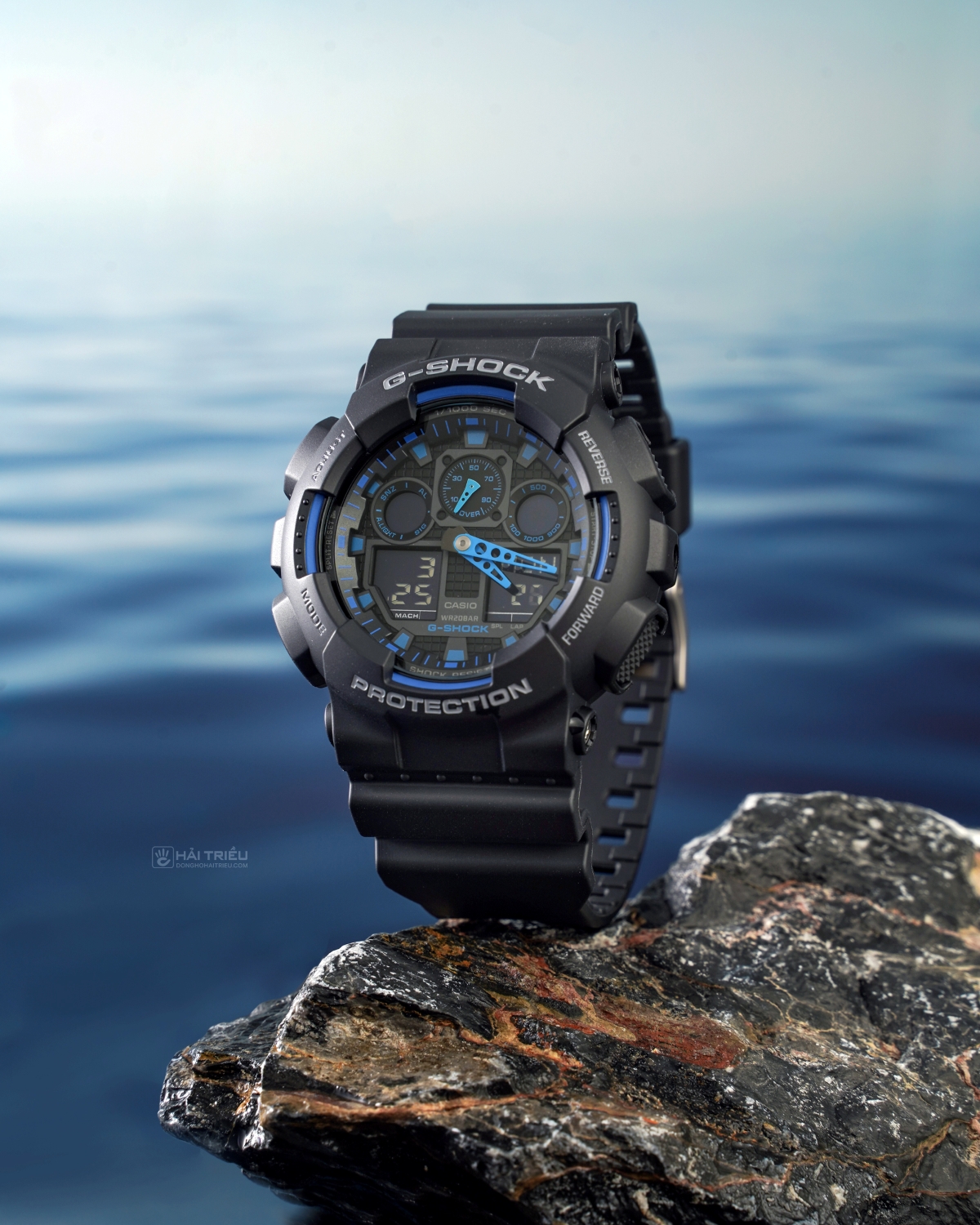 Đồng hồ G-Shock GA-100 màu đen xanh đặt trên đá cạnh mặt nước.