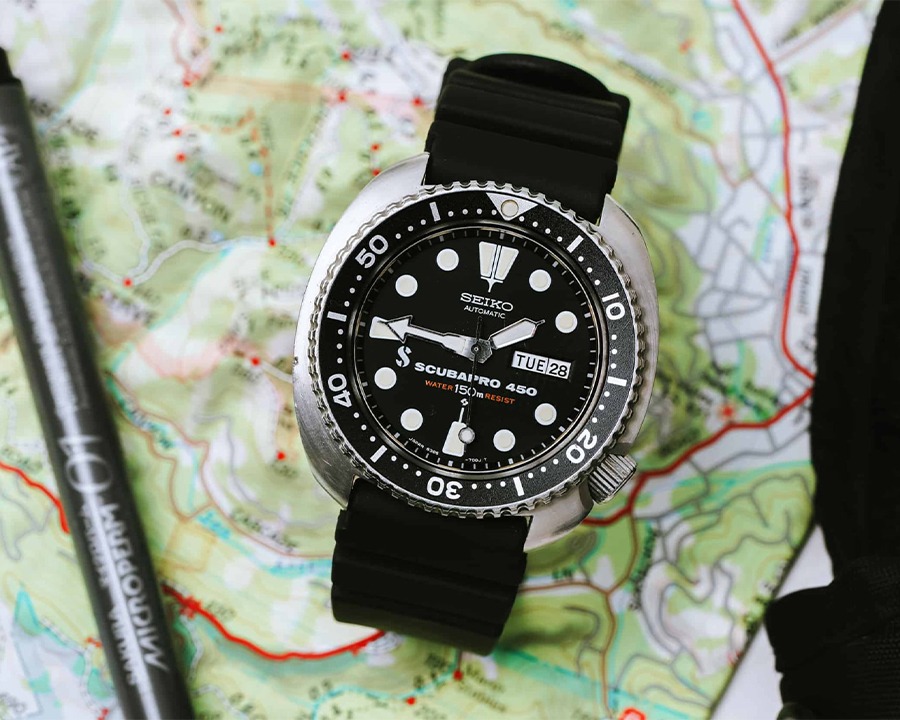 Đồng hồ lặn Seiko Turtle mặt số đen, vỏ thép dáng rùa kết hợp dây cao su, được đặt trên tấm bản đồ địa hình màu xanh lá.