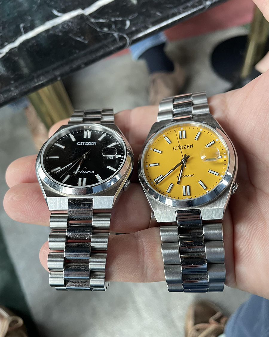 Hai chiếc đồng hồ Citizen Tsuyosa Automatic mặt số màu đen và màu vàng đặt cạnh nhau trên tay người dùng.