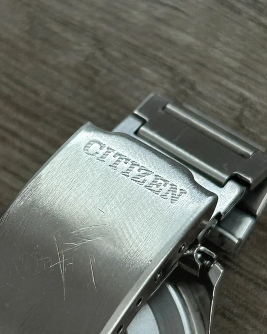 Khóa gập kim loại đồng hồ khắc logo Citizen có nhiều vết trầy xước trên bề mặt, đặt trên nền vân gỗ.