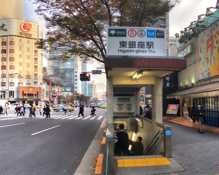 Cửa xuống ga tàu điện ngầm Higashi-ginza ở Tokyo. Bảng hiệu ga nổi bật phía trên lối đi bộ ngầm, kế bên là ngã tư đường phố có nhiều người qua lại.