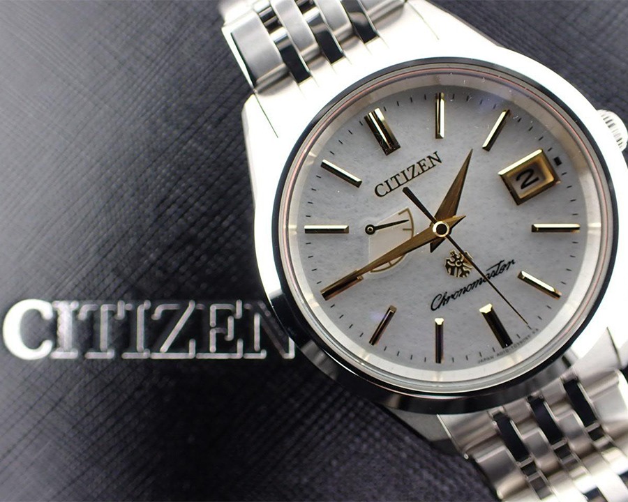 Đồng hồ Citizen Chronomaster mặt số vân giấy trắng, kim và cọc số vàng kim, dây đeo kim loại được đặt trên nền đen có nổi dòng chữ Citizen.