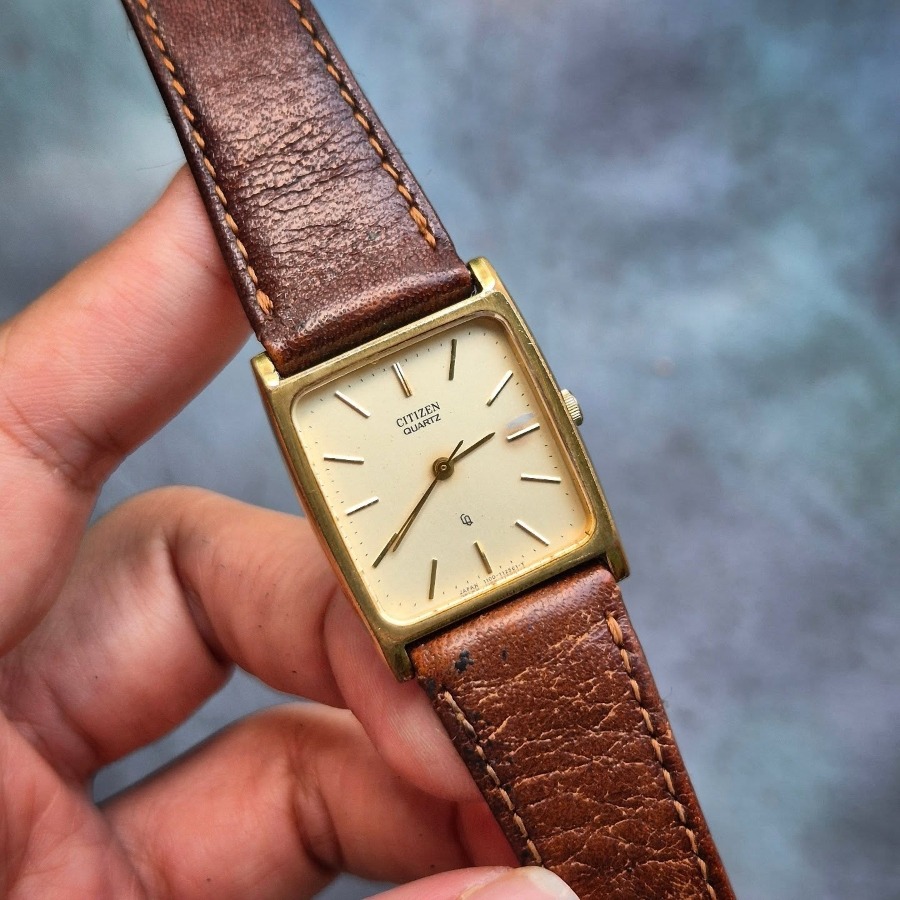 Cận cảnh tay cầm chiếc đồng hồ Citizen Quartz cổ mặt chữ nhật vỏ vàng với phần dây da màu nâu đã bong tróc và sờn cũ rõ rệt.